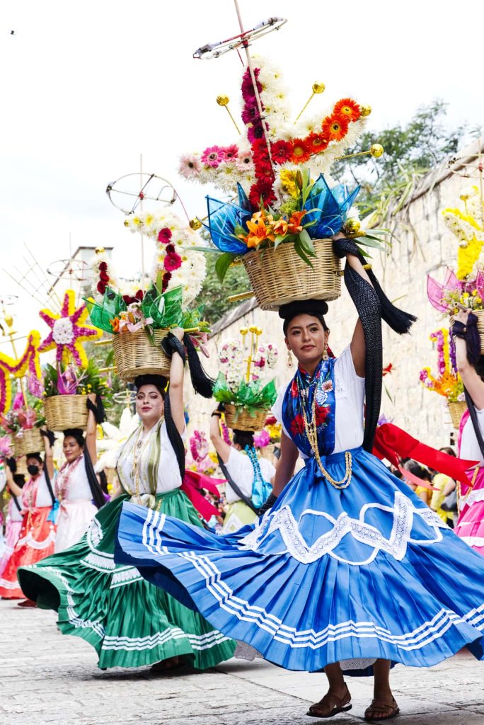 Las Chinas Oaxaqueñas dance in a line during a calenda in Oaxaca City during La Guelaguetza. The women each wear a different colored skirt and a matching scarf over a white shirt. A different, black scarf drapes behind them and up their arm which holds a basket with flower decorations and explosives.