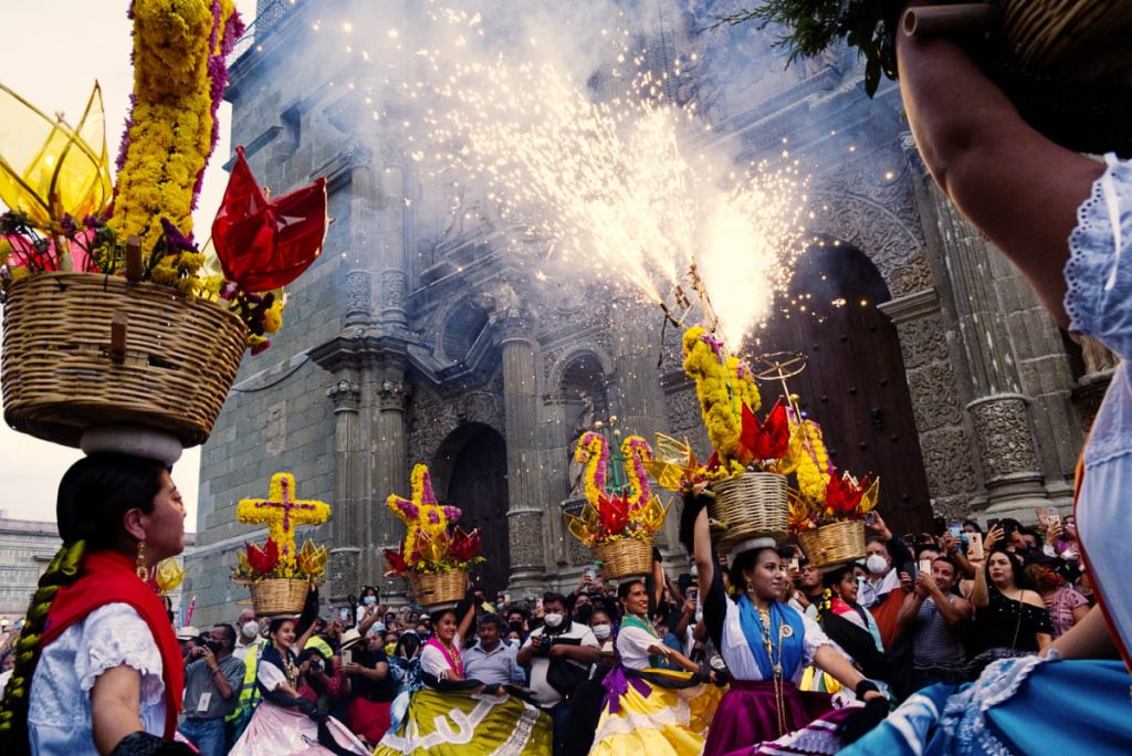 Chinas Oaxaqueñas in colorful skirts and scarves dance in front of the Oaxaca Cathedral at the end of a calenda, balancing flower-covered baskets shaped like crosses and stars, one shooting sparks into the air.