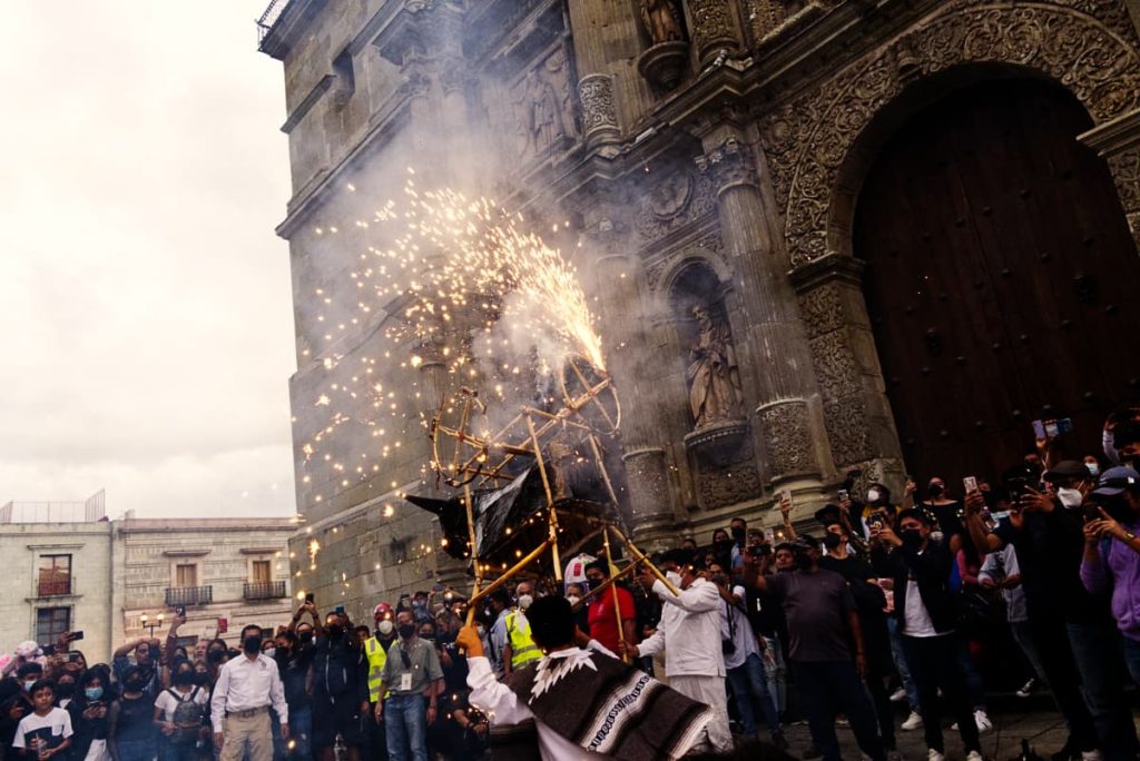 The back of a young boy swirling with El Torito, a bull-shaped bamboo frame rigged with fireworks, in front of the Oaxaca Cathedral at the end of a calenda, sparks bursting overhead as spectators crowd around