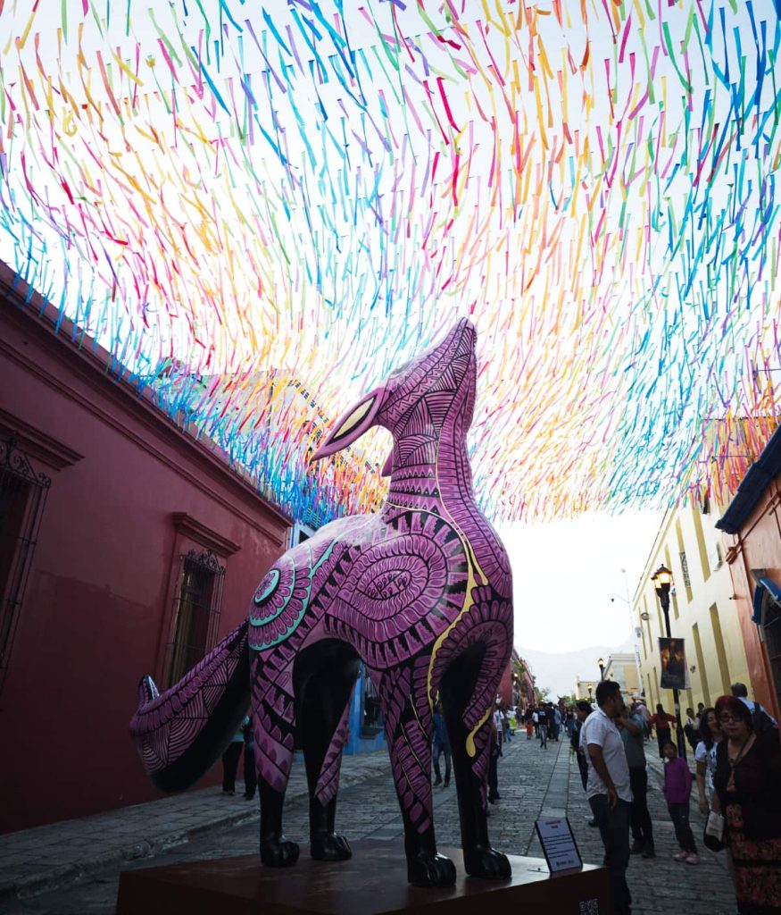 A street in Oaxaca with a large fox statue painted with pink and blue in the style of alebrijes. A few people mingle on the street under thin strips of colored papers strung across the street and fluttering in the wind.
