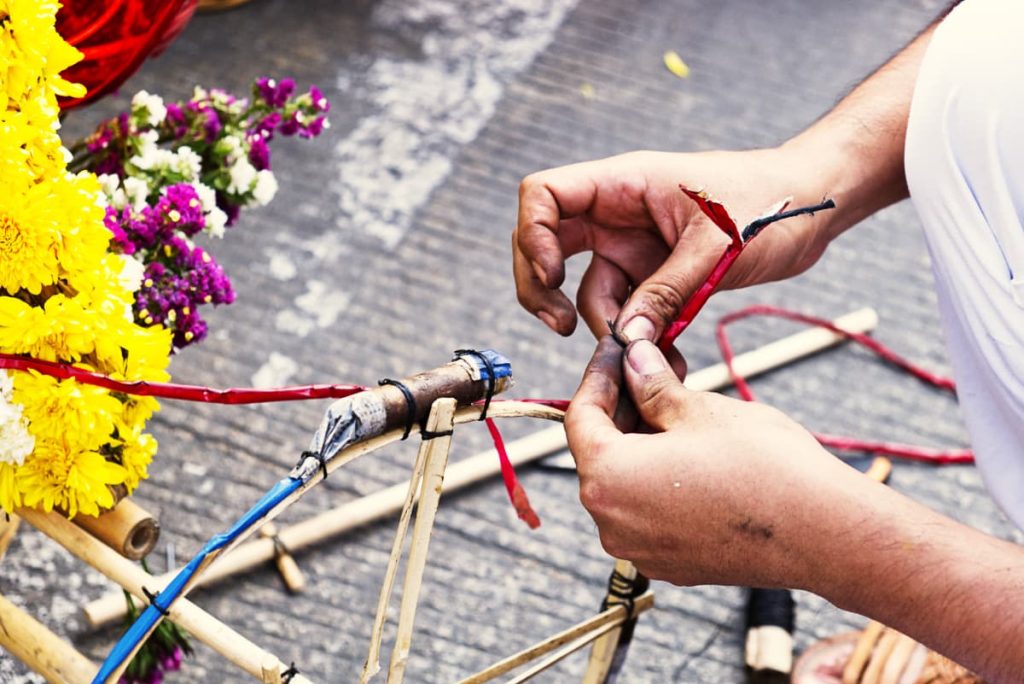 A closeup of hands covered in black soot as they fix a loose piece of explosives at a parade in Oaxaca. The fireworks are connected to a basket with bright yellow and purple flowers, which is partially visible on the left side of the frame.