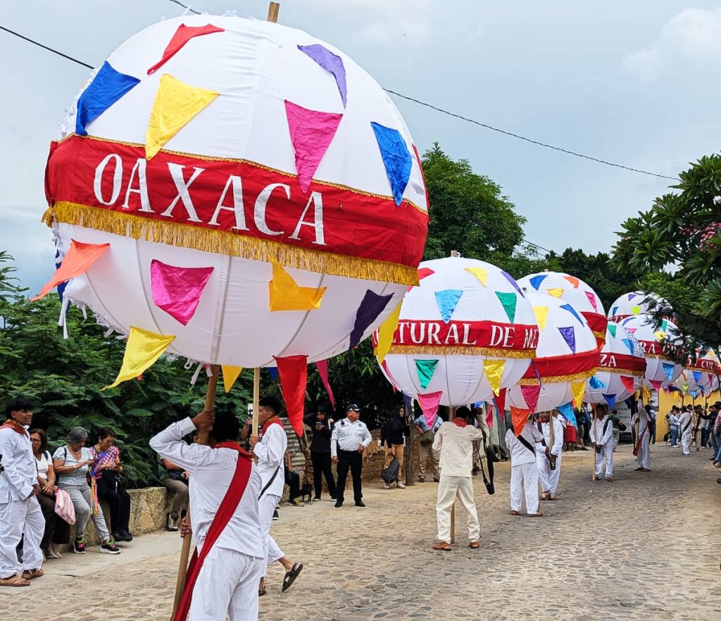 A line of men carry marmotas, white spherical lanterns on poles decorated with colorful triangular flags and a red ribbon reading 'Oaxaca,' down a cobblestone street during a calenda.