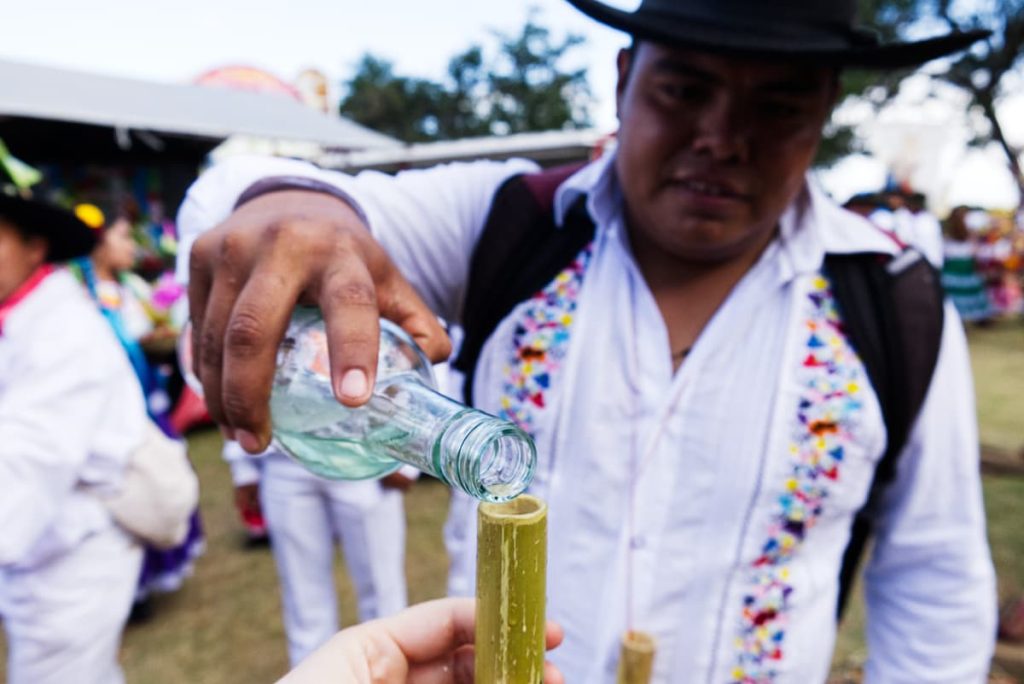 A blurred view of a man as he pours a shot of mezcal into a skinny piece of bamboo cut like a shot glass. He is wearing a white button down shirt with embroidered flowers on both sides and a black cowboy hat.