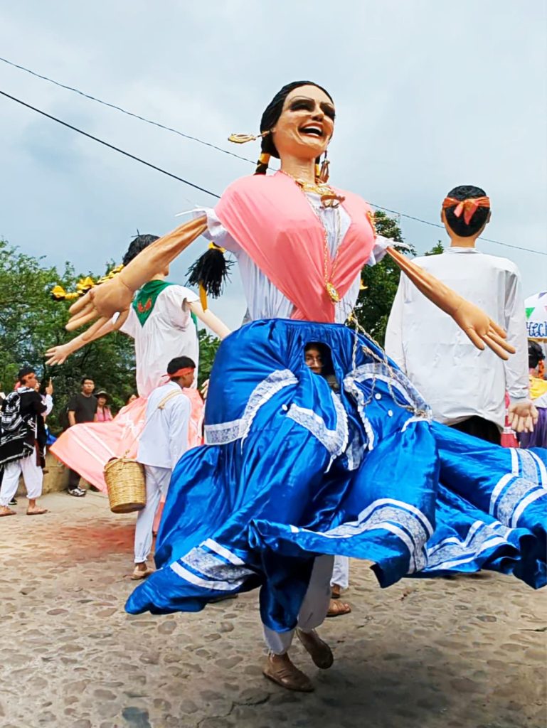 A female mono (puppet) spins at a parade in Oaxaca. She is larger than life-size and dressed in a traditional blue skirt, white top with a pink scarf, braids with ribbon, and earrings. Peeping out from below her waist is a man's face and at the bottom his legs. He controls the puppet during the calenda.