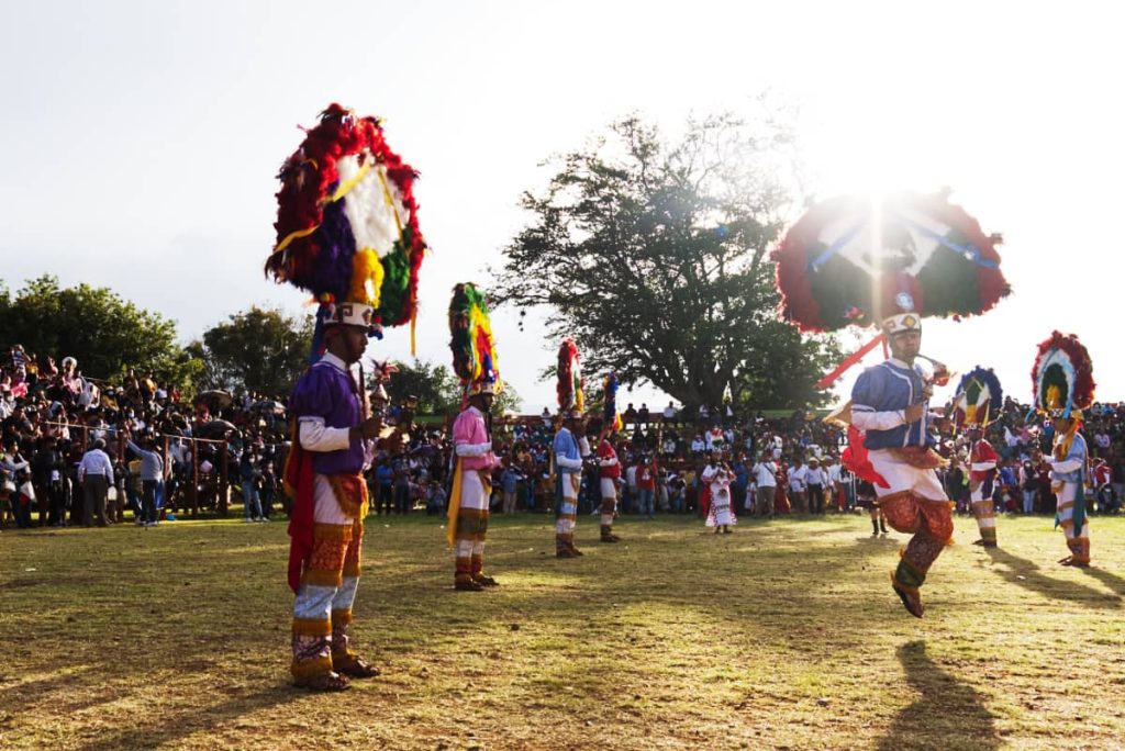 Young men wearing a feather headdress dance La Pluma de La Danza in Oaxaca on a grassy field surrounded by spectators.