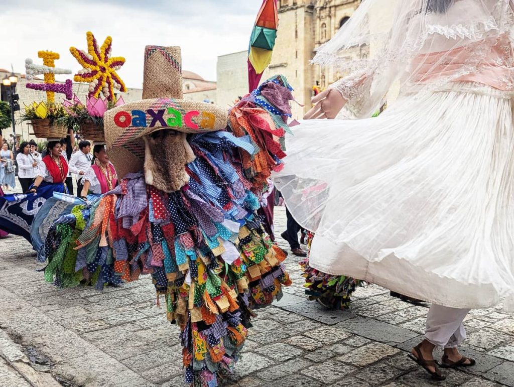 A tiliche dressed in a costume of colorful, cut up rags with a tall straw hat and animal fur covering their face dances in a wedding parade in Oaxaca in front of Santo Domingo church.