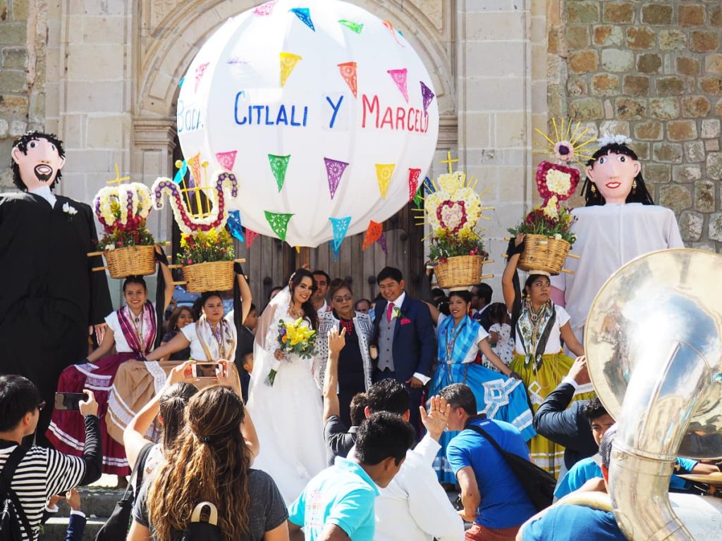 The bride and groom pose for photos in front of the church surrounded by different elements of a wedding parade in Oaxaca City. They include chinas Oaxaqueñas holding baskets of flowers, two large monos (puppets) that resemble the bride and groom. Behind them is a large marmota, that says Boda Citlali y Marcelo to announce the event.