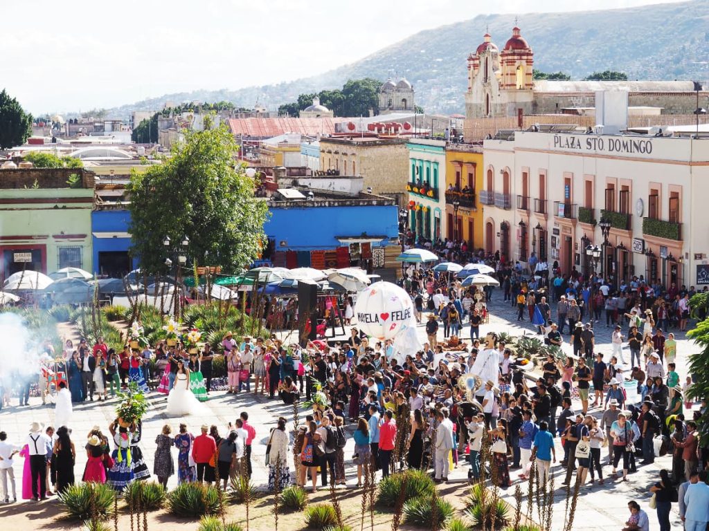 A wedding parade in Oaxaca seen from above, looking down at the square in front of Santo Domingo. The groom holds a bull structure lit with explosives while the bride stands near. They are surrounded by a crowd which includes two large mono puppets and a circular sphere with their names on it.