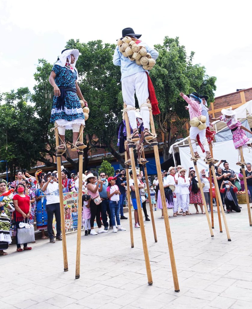 Young men stand on tall stilts in front of a crowd during a parade in Oaxaca. The Zancudos, as they are called, are wearing a hate and white pants, some with pastel colored tops and others with dresses.