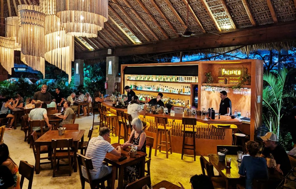 A view of the bar and dining room at El Nene seafood restaurant in Puerto Escondido. Under the palapa, macrame light fixtures hang above people sitting at tables in the dining room while bartenders make drinks at the bar.