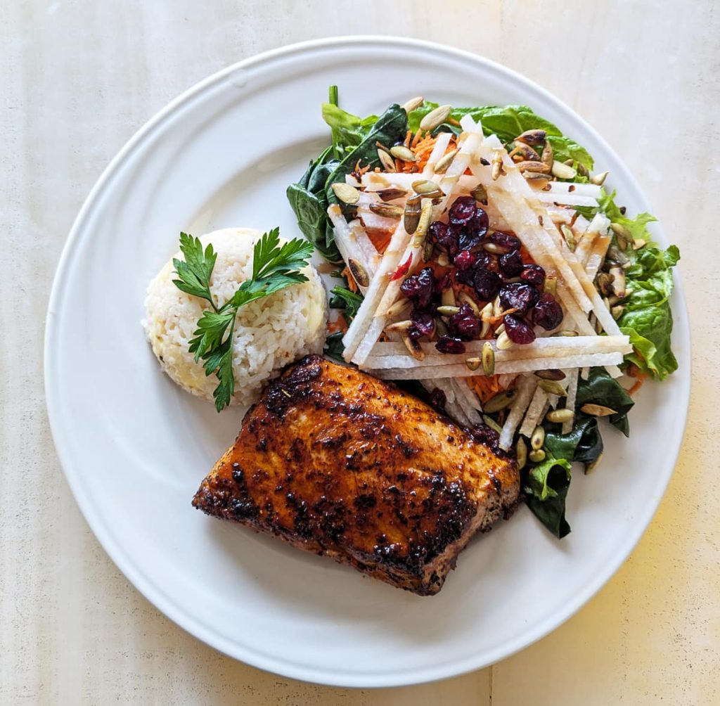 An overhead view of a white plate of seasoned grilled fish, rice, and a salad topped with jicama strips in a triangular shape, seeds, and cranberries.