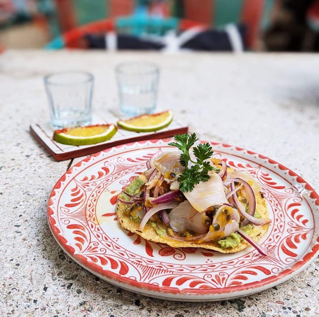 A white and red plate with a yellow corn tostada topped with maracuya ceviche at Maria Sirena seafood restaurant in La Punta. Behind the plate are two mezcal glasses, each with an orange dusted in red worm salt.