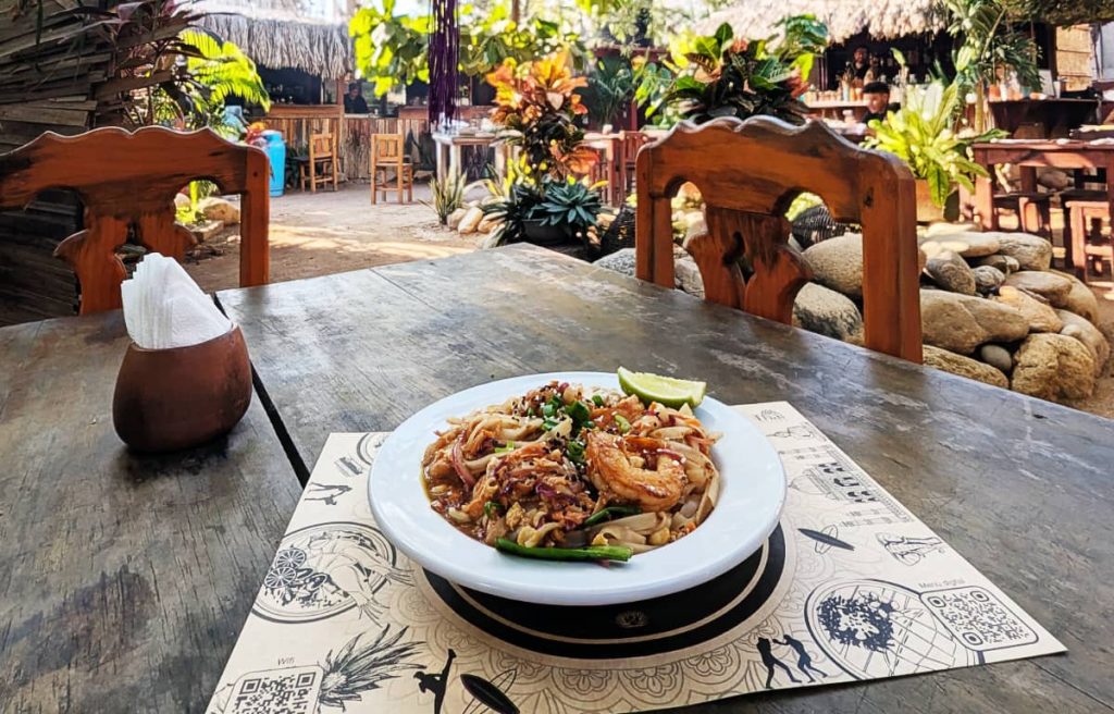 A shallow white bowl of pad thai with large shrimp and a lime wedge on a wooden table in an outdoor garden area. In the background is a palapa bar and tropical plants.